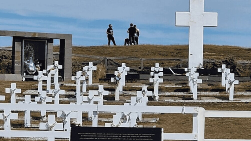 A cemetery with numerous white crosses, a large central cross, and two people walking in the background. 1