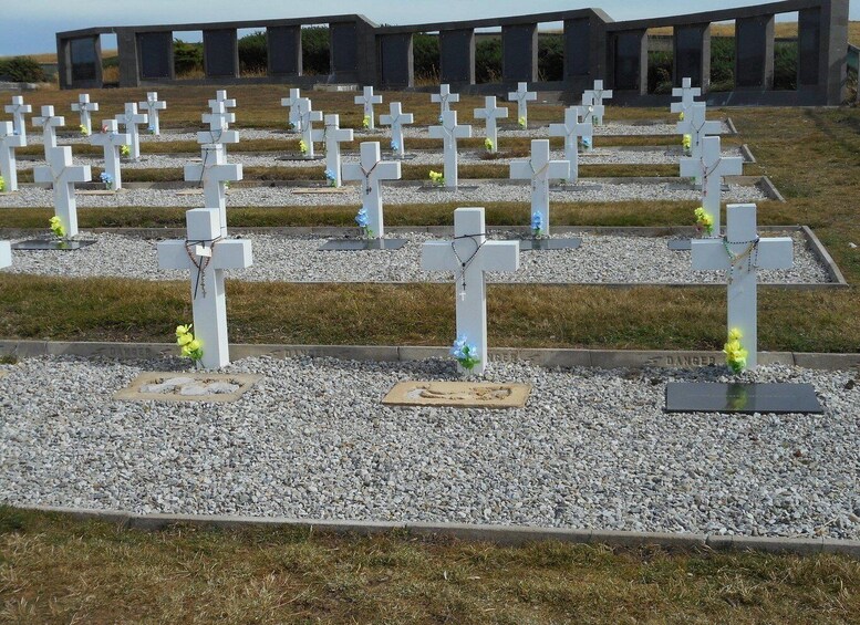 A cemetery with numerous white crosses, some adorned with flowers, set against a backdrop of a modern building. 2