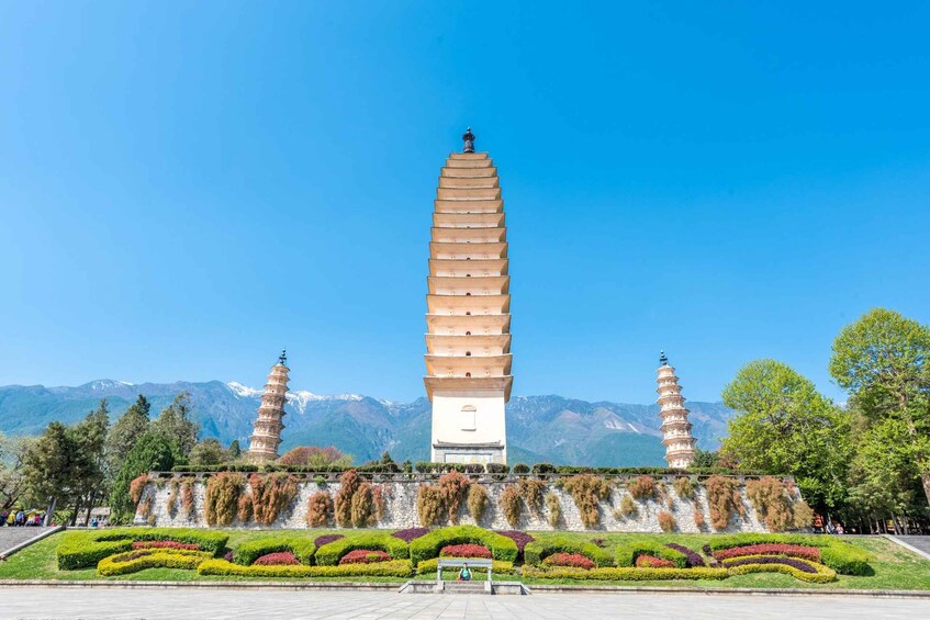 A large pagoda with a white base and multiple tiers, surrounded by a landscaped garden with colorful flower beds, set against a backdrop of mountains and clear blue sky. 1