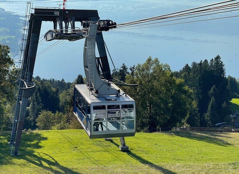 A cable car suspended by cables, a grassy hill, and a forested area with a body of water in the background. 1
