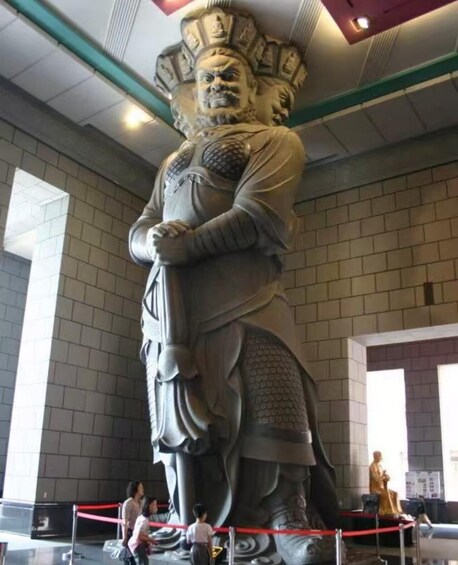 A large statue with intricate details, including a headdress and armor, standing in a spacious indoor area with tiled walls and a high ceiling. 2