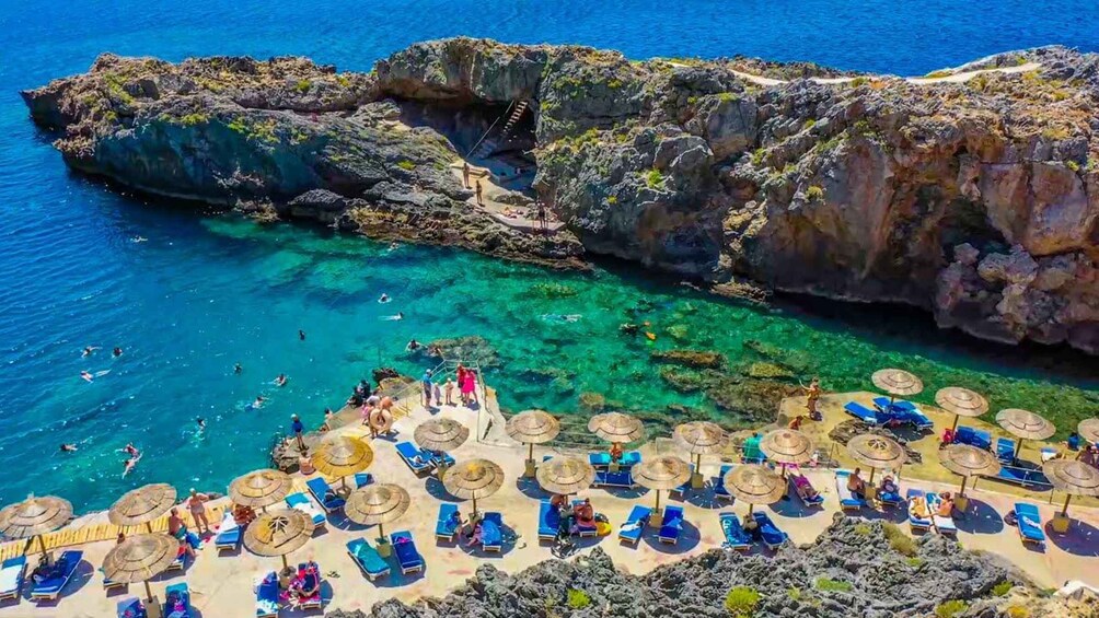 A coastal area with clear blue waters, rocky cliffs, and a beach with sun loungers and umbrellas. 1