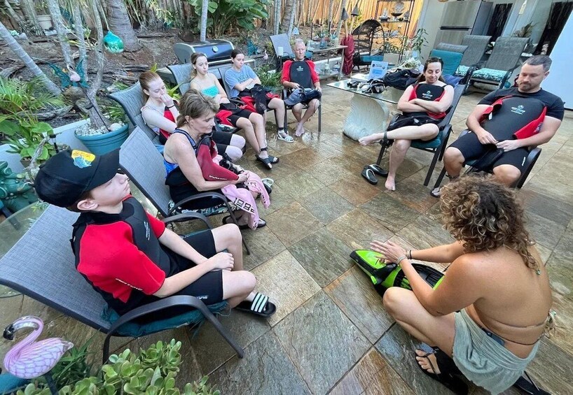 A group of people sitting on chairs outdoors, some wearing life vests, with a pool and plants in the background. 5
