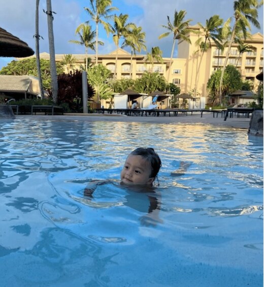A child swimming in a pool with palm trees and a hotel in the background. 1