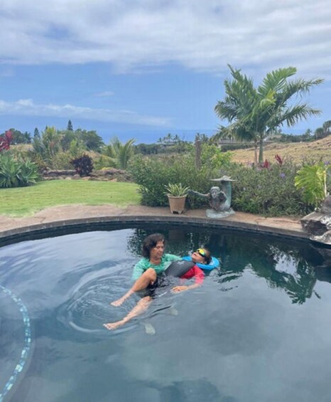 A person in a swimming pool with a child, surrounded by tropical plants and a clear sky. 4