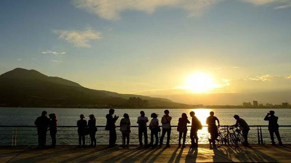 A group of people silhouetted against a sunset by a body of water with mountains in the background. 3