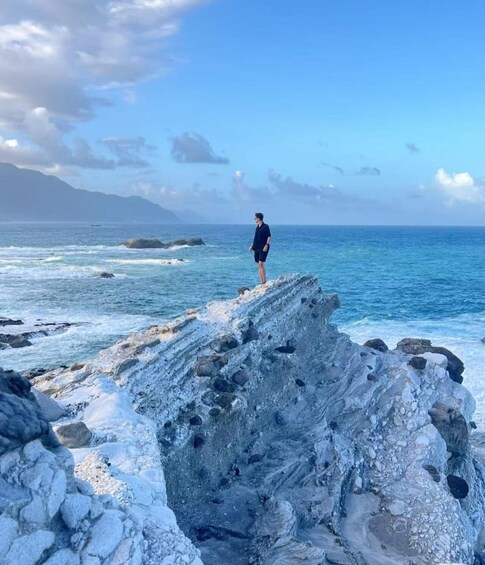 A person standing on a rocky outcrop by the sea with waves crashing below. 5