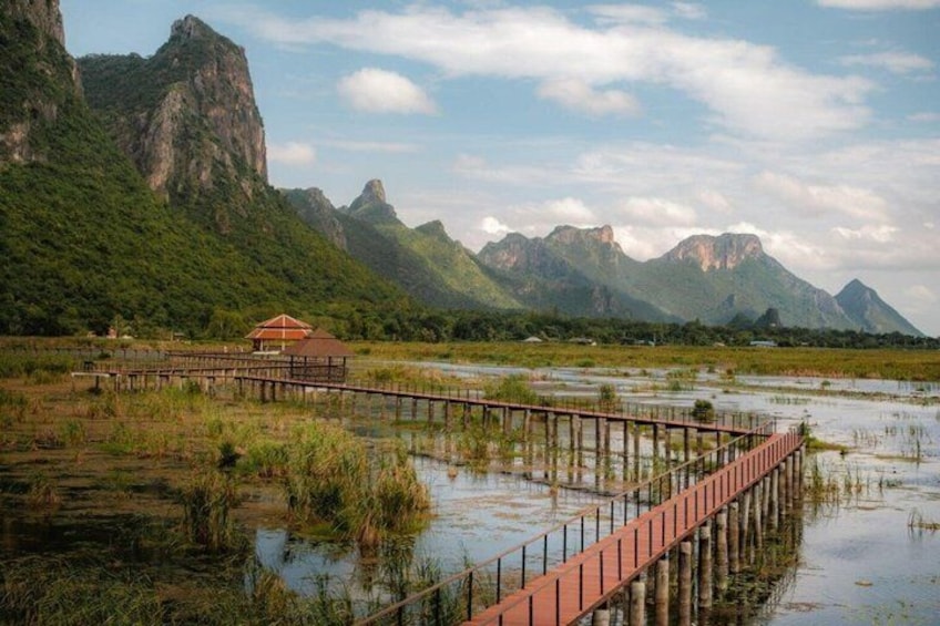 A wooden bridge over a marshy area with a small building on the left and mountains in the background. 1