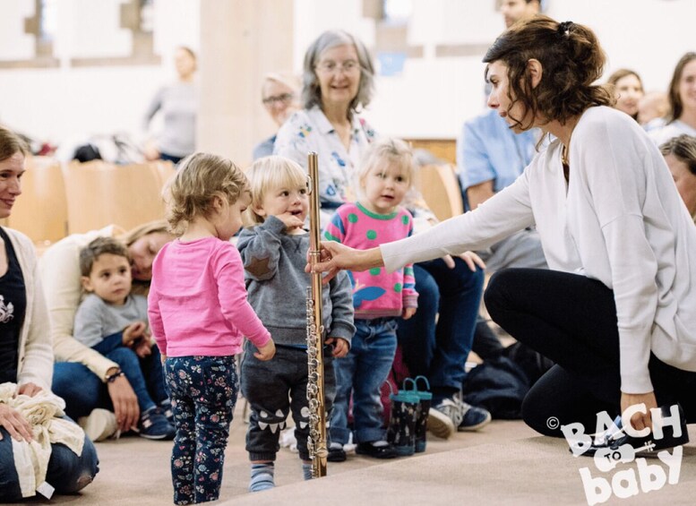 A group of people, including children, gathered around a musical instrument, possibly a flute, in an indoor setting with chairs and a carpeted floor. 5