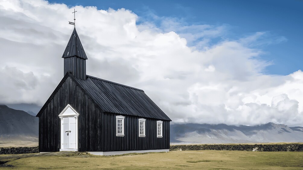 A black church with a white door and windows, a steeple with a cross, and a stone wall in a grassy field with mountains in the background. 4
