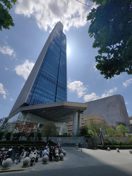 A modern skyscraper with a glass facade, surrounded by trees and a parking area with motorcycles. 3
