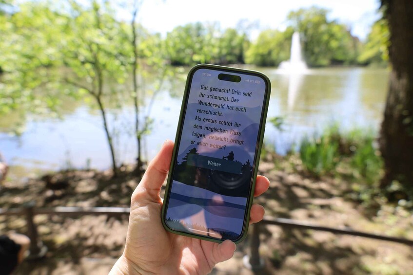 A hand holding a smartphone displaying text in German, with a blurred background of a lake and trees. 5