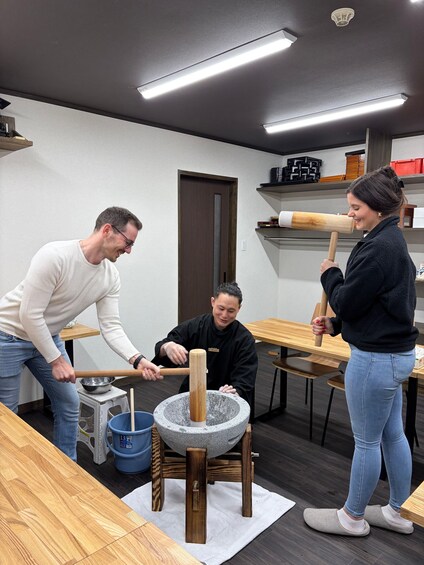 Three people using a mortar and pestle, a wooden table, and a blue bucket in a room with shelves and a door. 2