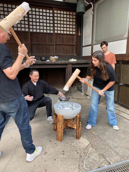 People engaged in a traditional food preparation activity outdoors, using wooden tools and a stone mortar. 4