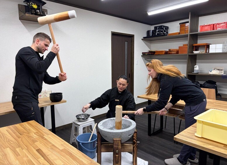A person using a mortar and pestle, another person holding a wooden mallet, and a third person observing. 1