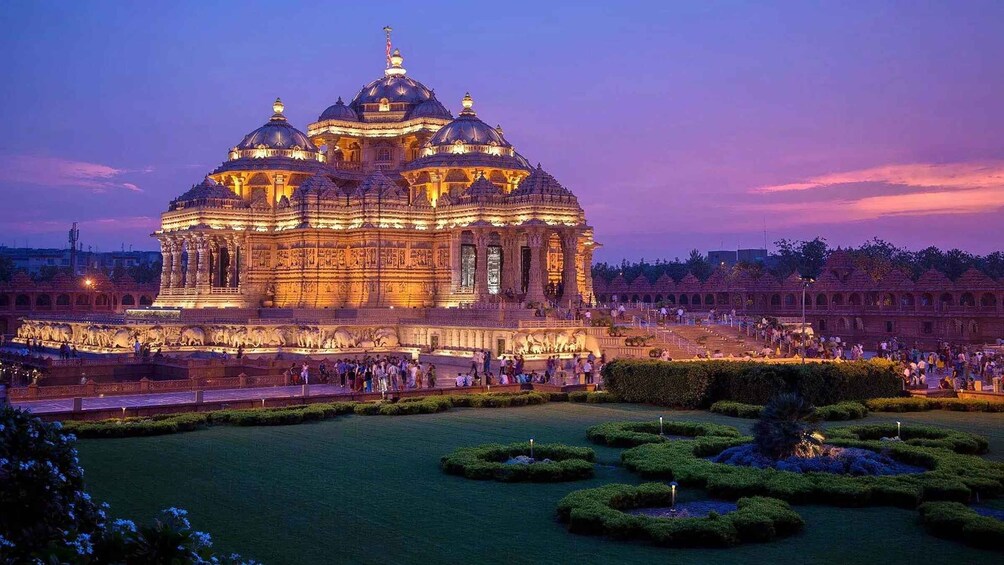 A large, illuminated temple complex with multiple domes and intricate carvings, surrounded by a well-maintained garden and a crowd of visitors. 2