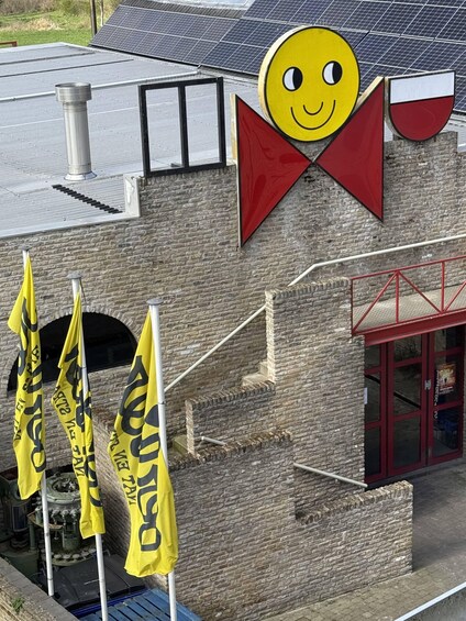 A building with a brick facade, solar panels on the roof, a large red and white sign, and yellow flags with black text. 3