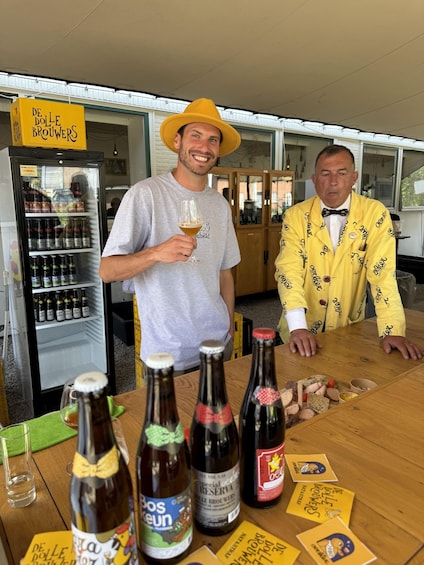Two men standing at a table with bottles and a refrigerator in the background. 4