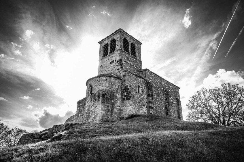 A historic stone church with a bell tower, surrounded by a grassy hill and trees under a cloudy sky. 1
