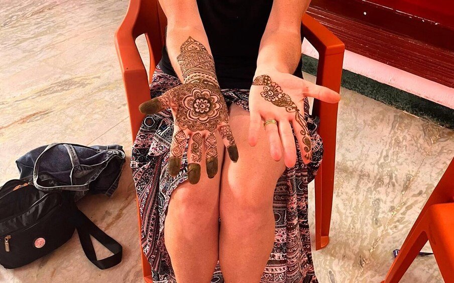 A person with intricate henna designs on their hands and legs, seated on a chair with a patterned skirt. 1