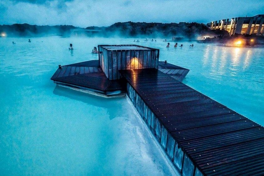 A wooden dock extending into a geothermal spa with people swimming and a building with a light on it. 1
