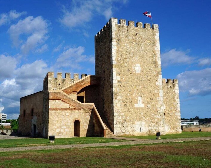 Eine historische Steinburg mit einer Flagge auf dem Turm, einem großen Bogenportal und einem grünen Innenhof. 1