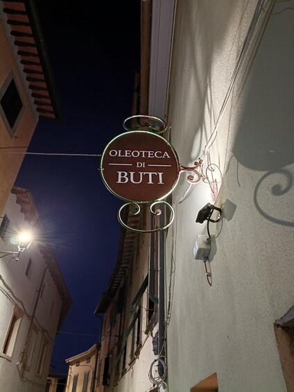 A street sign with the text 'OLEOTECIA DI BUTI' against a twilight sky. 5