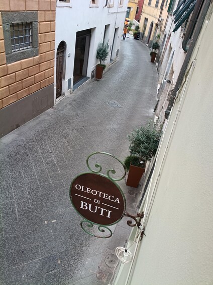 A narrow street with buildings on either side, a sign reading 'OLEOTECIA DI BUTI', and potted plants along the sidewalk. 2