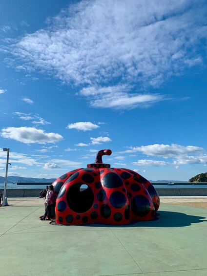 A large, red, polka-dotted structure resembling a ladybug with a tunnel entrance, located on a concrete platform by a body of water with mountains in the background. 1