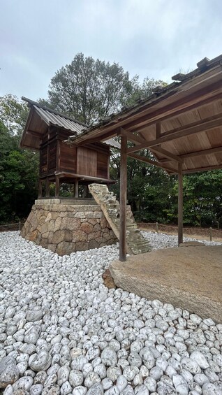 A traditional wooden structure with a stone base, surrounded by white pebbles and greenery. 3