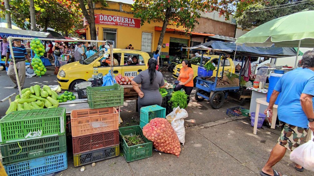 A street market with vendors selling fruits and vegetables, a yellow taxi, and a building with a sign that reads 'DROGUERIA PANAMERICANA'. 2