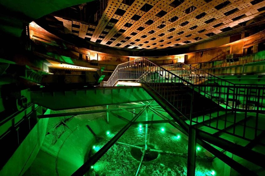 A dimly lit industrial interior with a staircase leading to a lower level, green lighting, and a metal ceiling with a grid pattern. 5