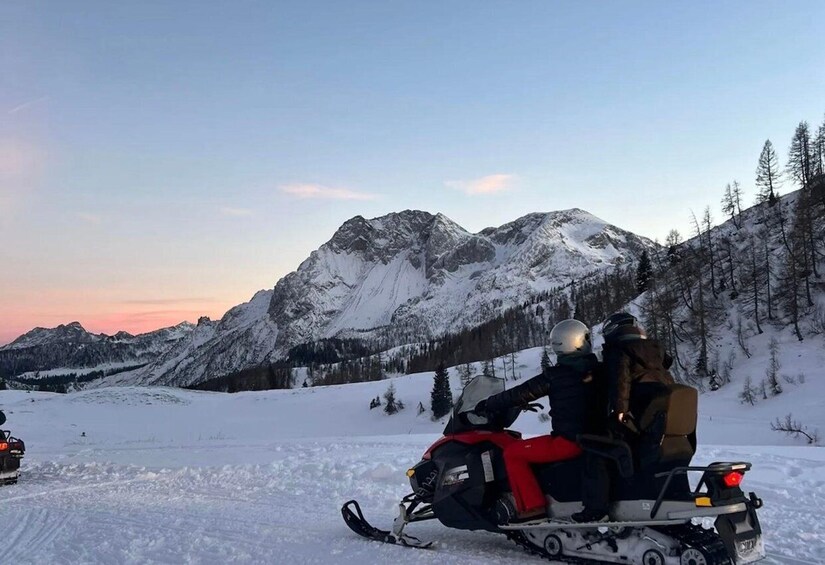 A snowmobile on a snowy mountain trail with a person riding it, surrounded by snow-covered peaks and trees. 5