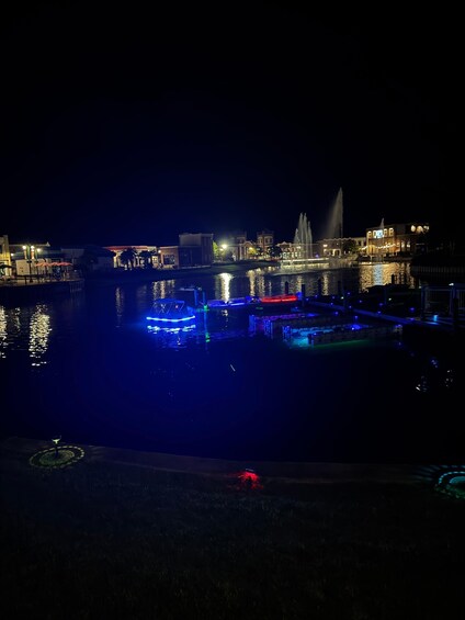 A nighttime waterfront scene with illuminated buildings, a fountain, and a dock with boats. 4