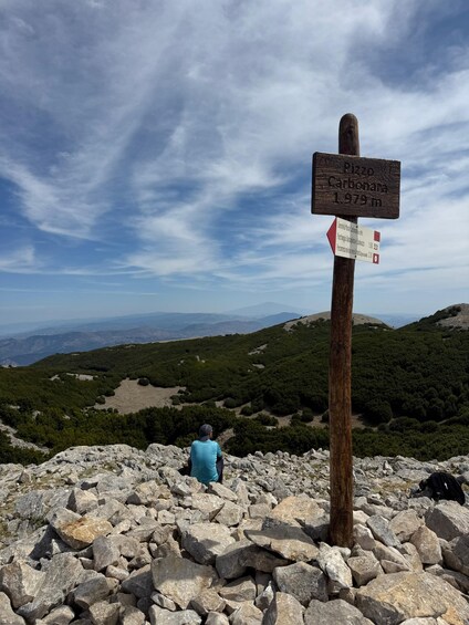 A rocky mountain landscape with a person sitting on a rock, a wooden signpost with route information, and a clear blue sky. 1