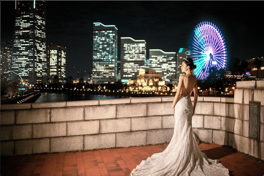 A person in a wedding dress standing on a bridge with a city skyline and a Ferris wheel in the background. 3