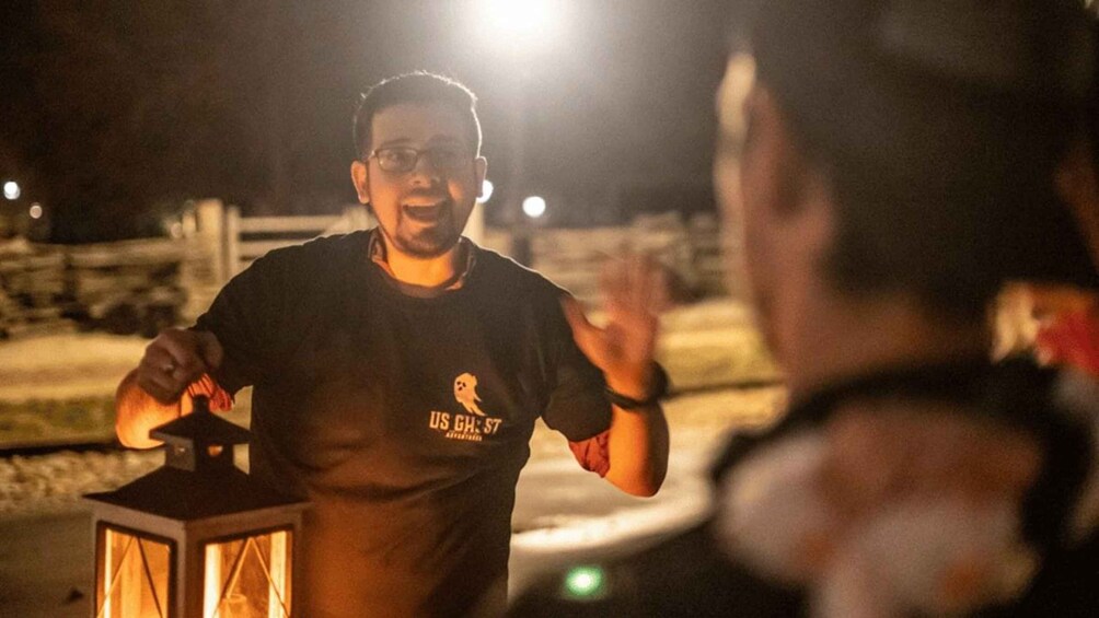 A person wearing a t-shirt with a ghost logo, standing outdoors at night with a lantern. 4