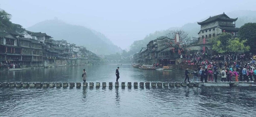 A misty river with traditional buildings on both sides, a person standing in the water, and a crowd on the right bank. 3