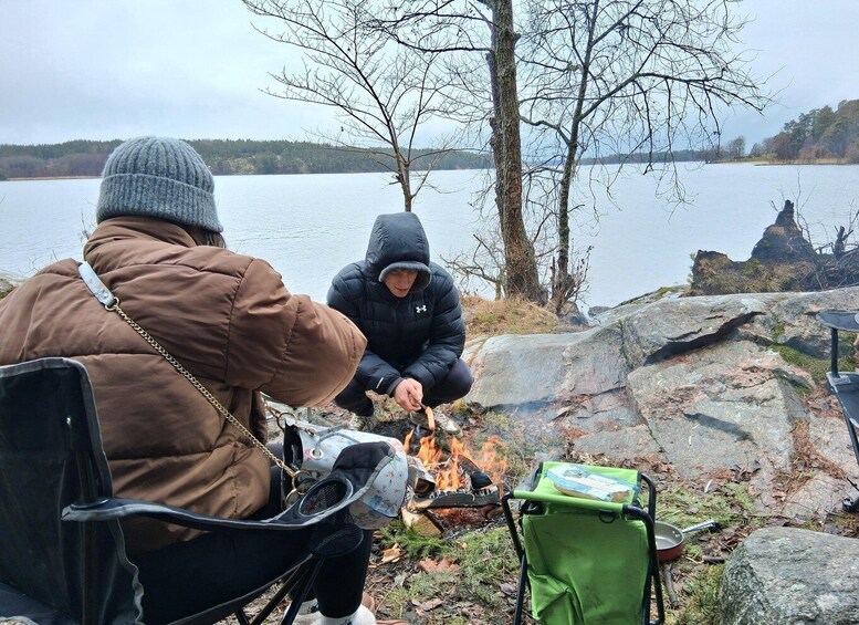 Two people sitting by a campfire near a lake, with one person cooking and the other holding a camera. 2