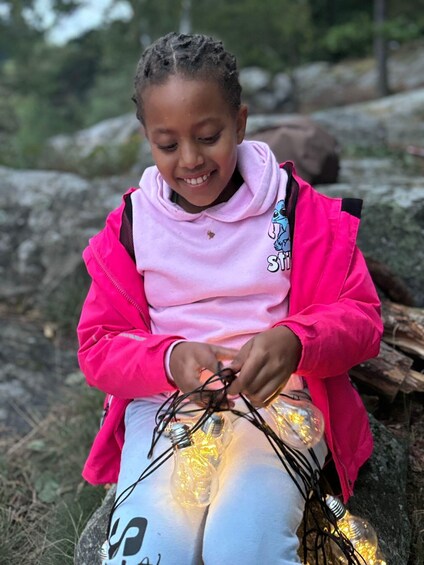 A child sitting outdoors, wearing a pink shirt and a bright pink jacket, with a backpack. 3