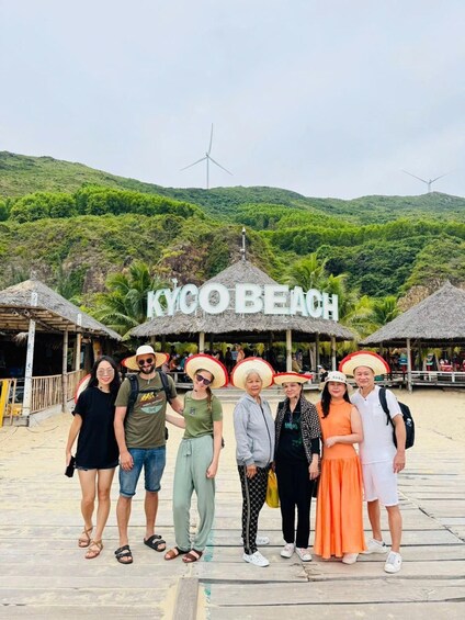 A group of people posing in front of a beach with a sign that reads 'KYCO BEACH' and wind turbines in the background. 2