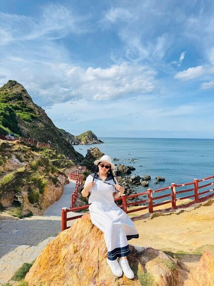 A person sitting on a rock by a coastal path with a view of the sea and a mountainous landscape. 4