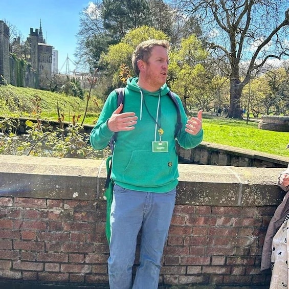 A person in a green hoodie standing by a brick wall with a park and a building in the background. 4