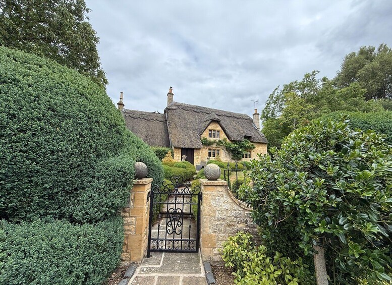 A traditional stone house with a thatched roof, surrounded by lush greenery and a gated entrance. 1