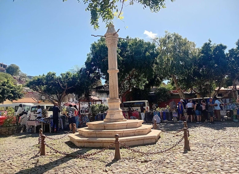 A stone monument with a statue, surrounded by a cobblestone area with chains, and a crowd of people in the background. 3