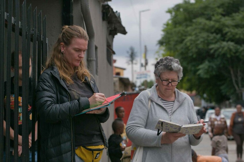 Two women, one holding a clipboard and the other holding a book, standing on a street with a fence and buildings in the background. 3