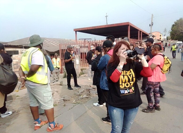 A street scene with people, some wearing safety vests, and a woman taking a photo. 1