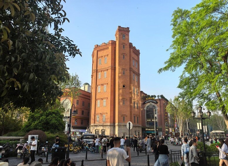 A busy urban square with a prominent red brick tower, surrounded by trees and various shops. 3