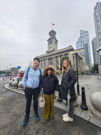 Three people standing in front of a historic building with a clock tower, in an urban setting with modern skyscrapers in the background. 1