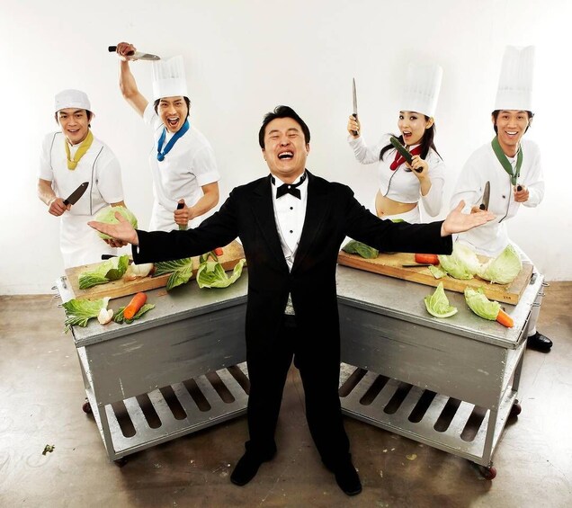 A man in a tuxedo with arms outstretched, surrounded by four chefs holding knives and cutting vegetables. 5